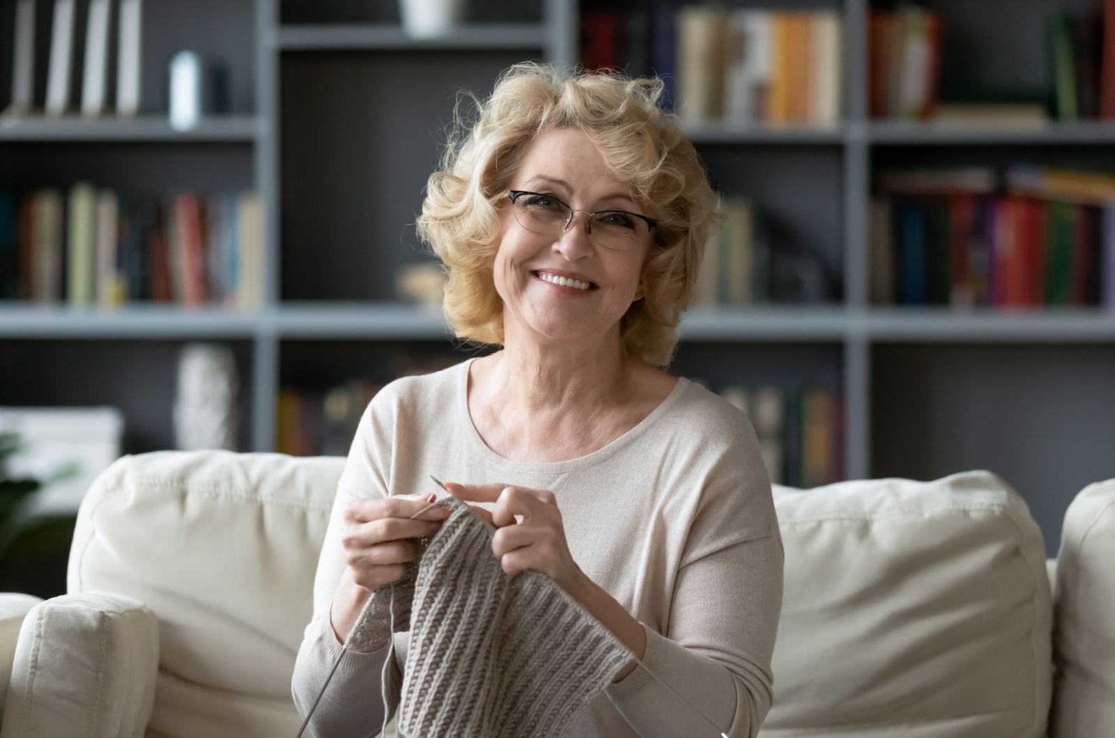 A smiling older adult sitting on the couch and knitting.