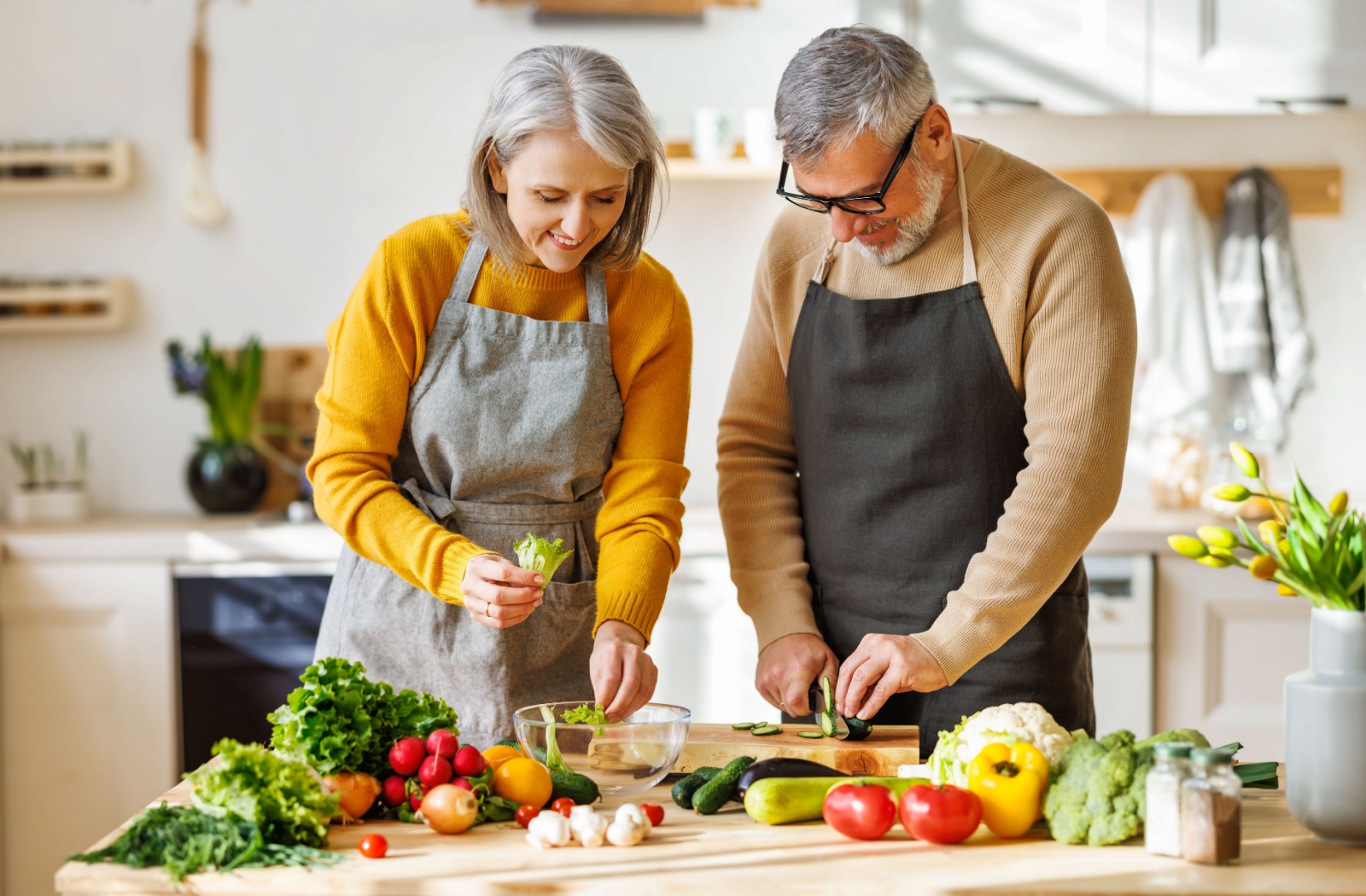 An older couple in a bright kitchen chopping and preparing a variety of colorful vegetables.