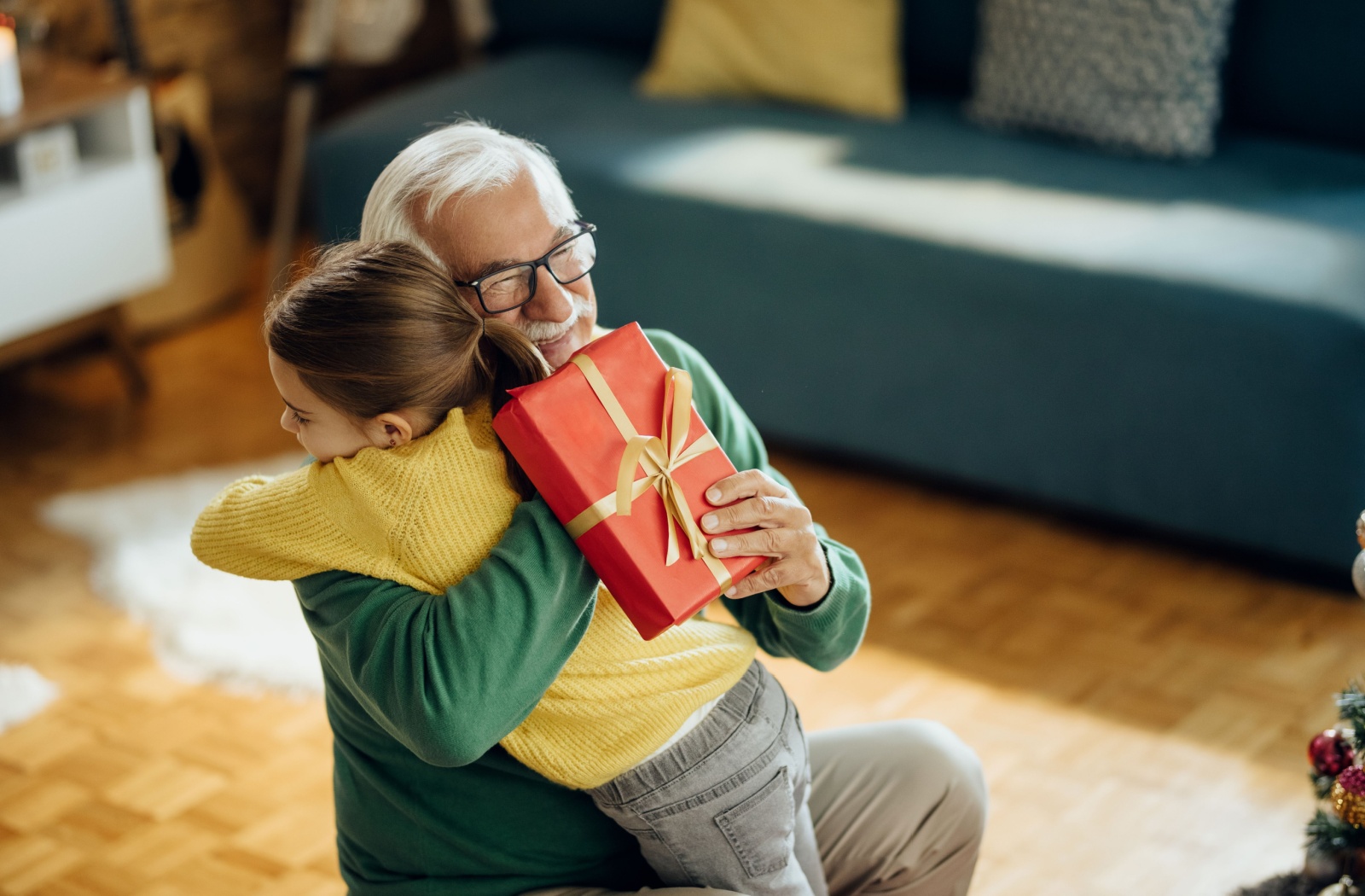 A grandparent smiling and hugging their grandchild while accepting a gift wrapped in red paper with gold ribbon.