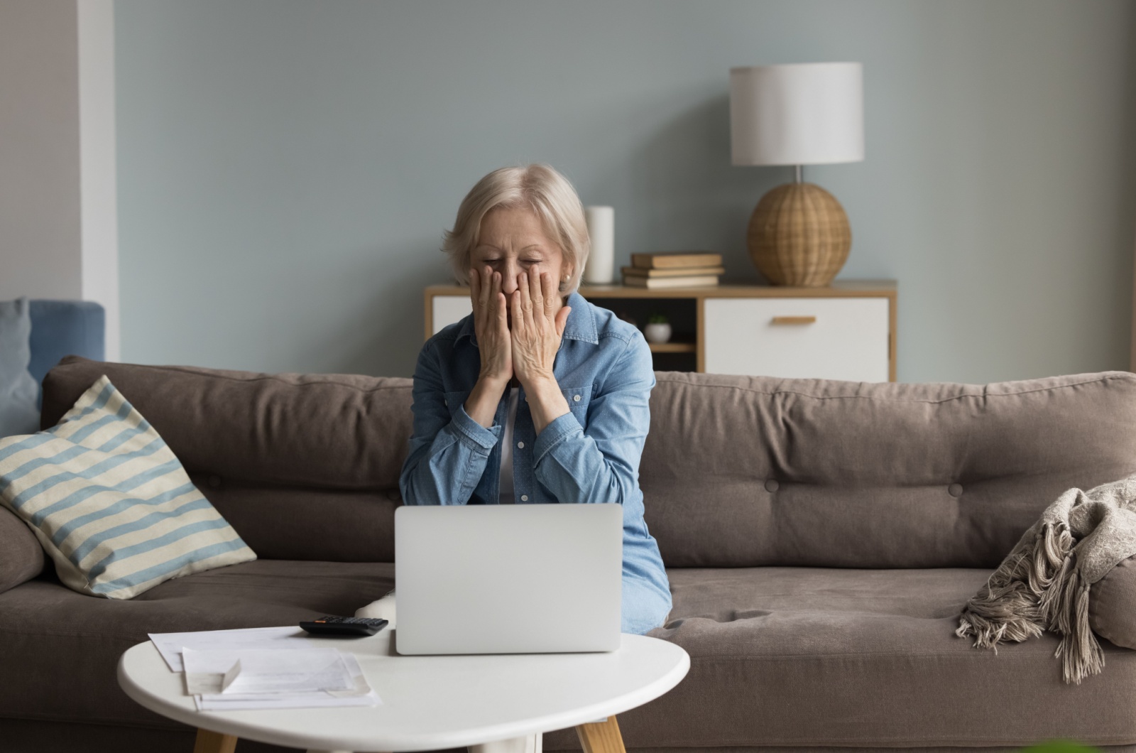 An older adult sitting in front of their laptop with their hands on their face in shock from an online scam.