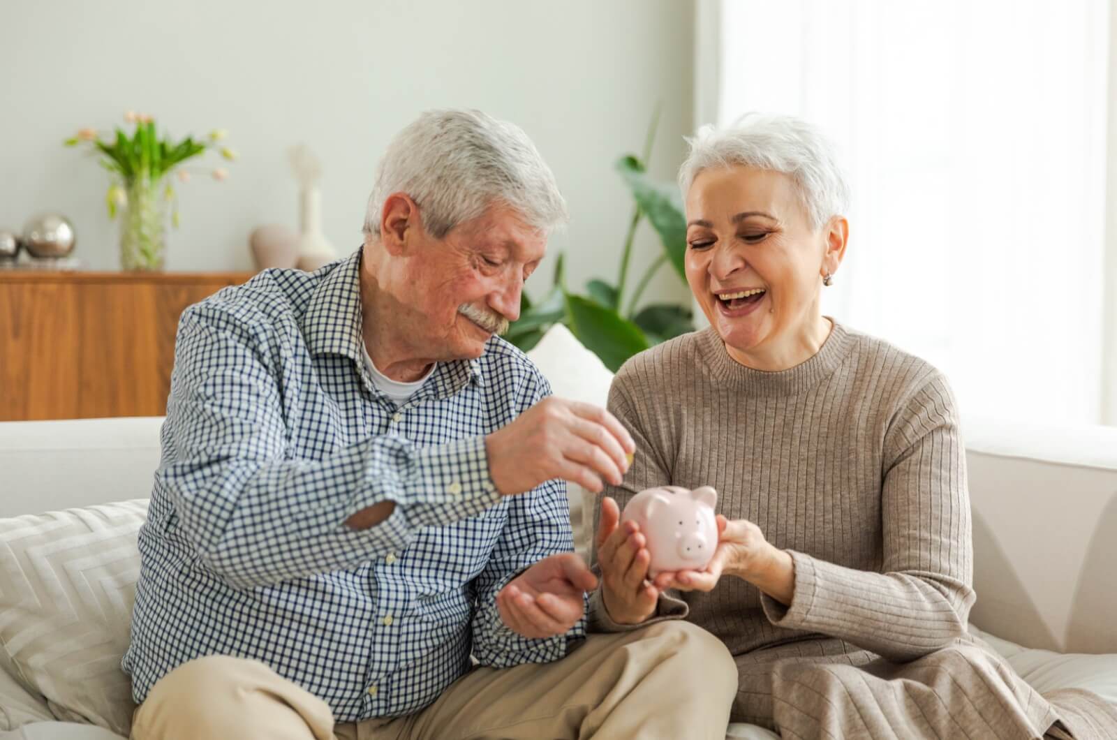 A happy older couple sitting on a couch putting coins in a piggy bank.